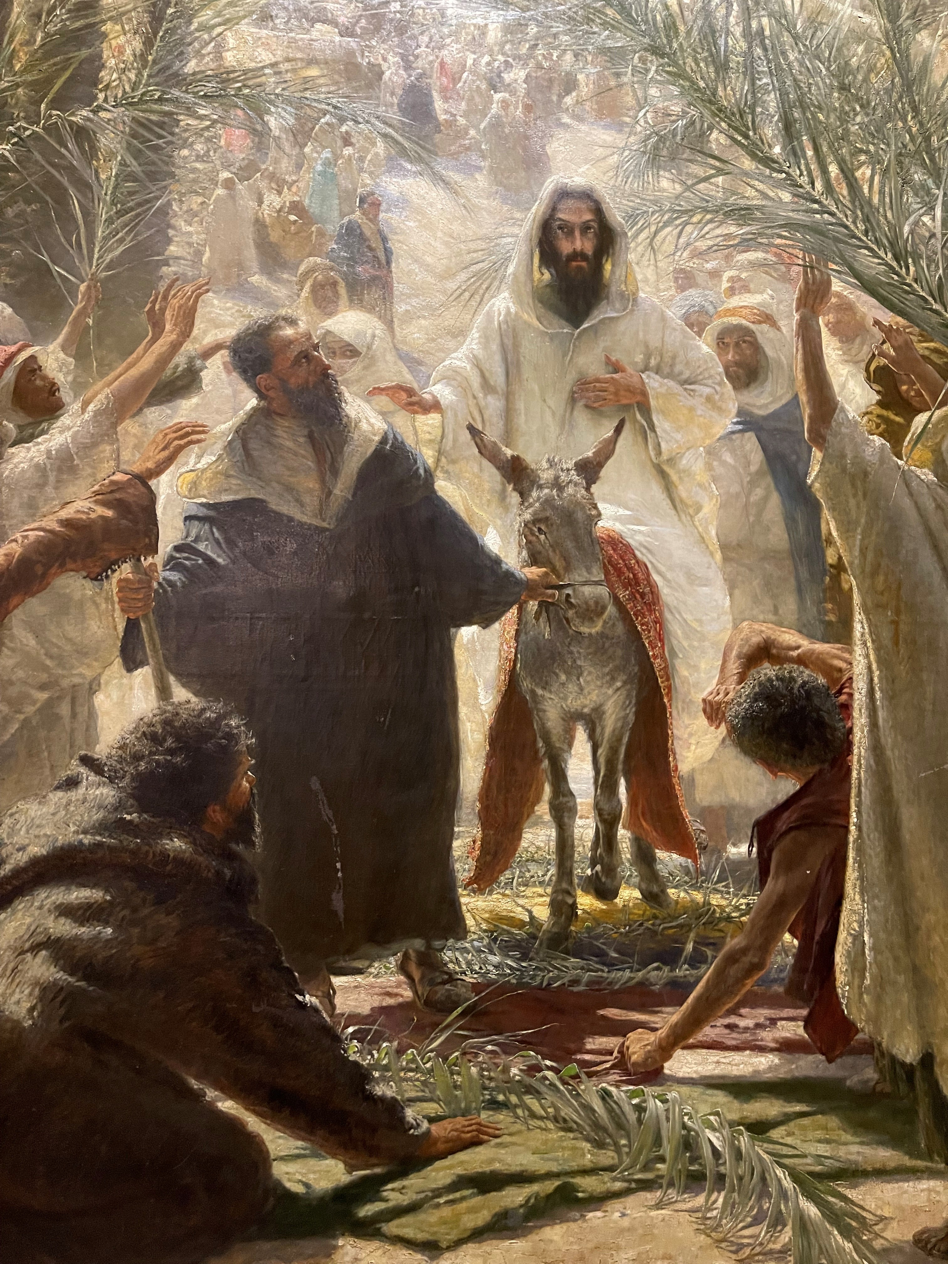 Jeesus ratsastaa aasilla palmusunnuntaina. Ihmiset levittävät maahan palmunoksia.